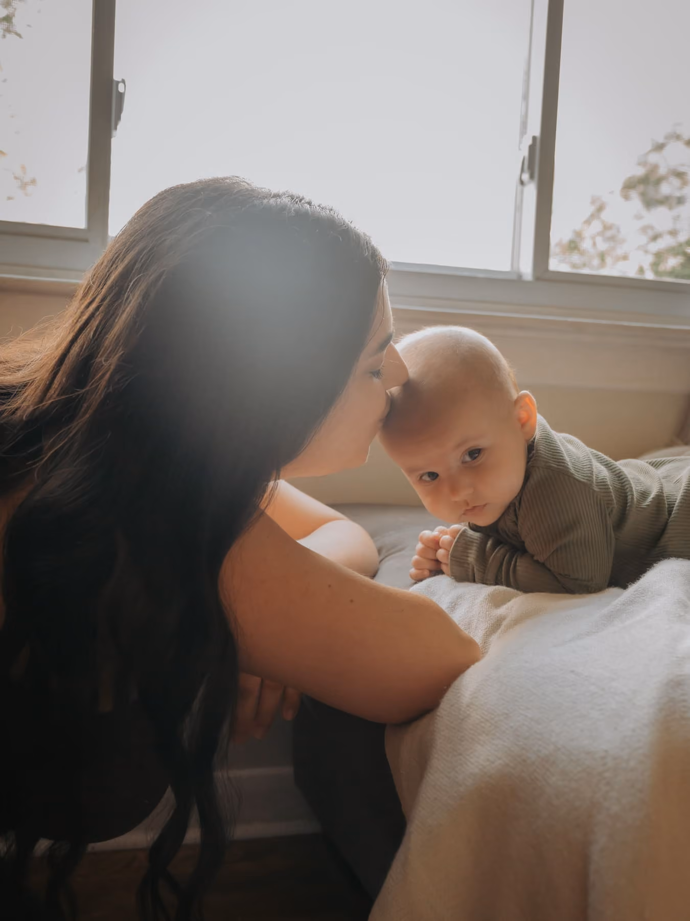 A mother kisses her baby’s head on the bed, in window light.
