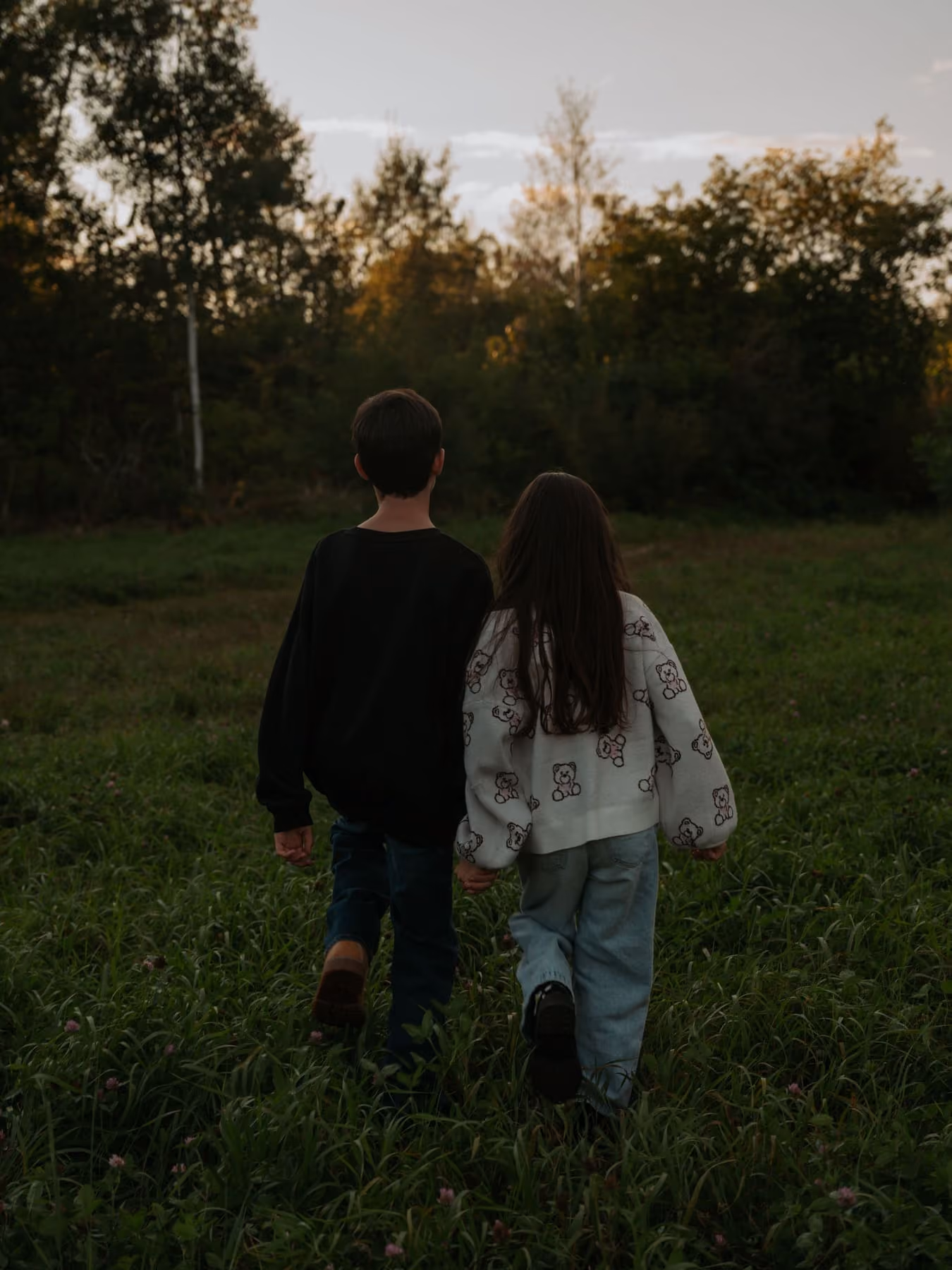 Two children walk away across the long grass at dusk.
